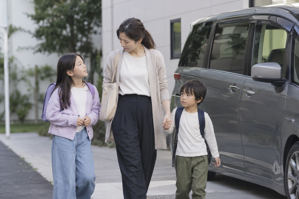送迎の車のそばを二人の子どもと歩く日本人の母親